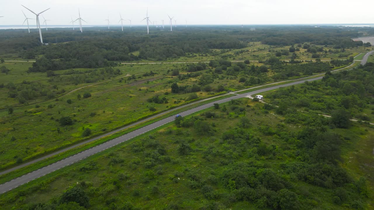 Aerial drone footage flying over a cloudy and misty peninsula where a small blue car drives on a rural countryside road into the distance in front of large white wind turbines at a wind farm, Paldiski
