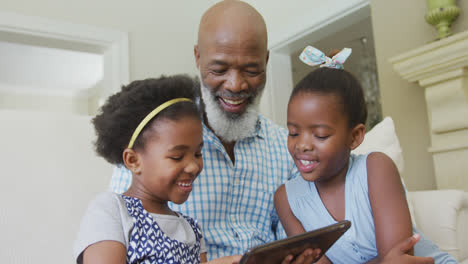 Feliz-Abuelo-Afroamericano-Con-Nietas-Usando-Tableta-En-La-Sala-De-Estar