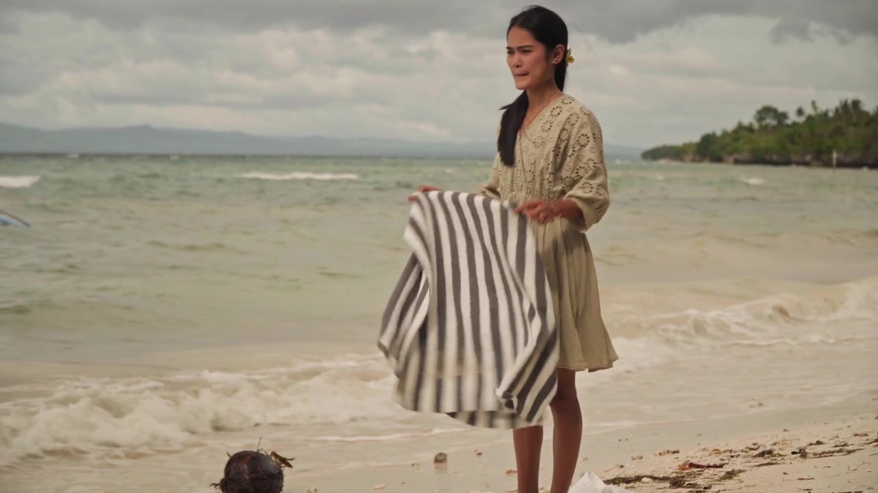 Mulher asiática na praia dobrando a toalha e arrumando a sacola, orla arenosa com casca de coco, ondas suaves quebrando, céu nublado, cabelo trançado, vestido vintage, humor tranquilo e reflexivo, solidão à beira-mar, em um ritmo lento.