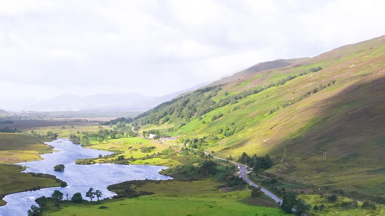 Drone pans above winding river, lush green meadows, and hills in bright natural daylight