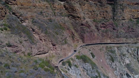 Toma-Panorámica-De-Un-Dron-De-Un-Coche-Conduciendo-Por-Una-Pintoresca-Carretera-Costera-Cortada-En-Un-Acantilado-Junto-Al-Océano---Chapman&#39;s-Peak-Drive,-Península-Del-Cabo,-Sudáfrica