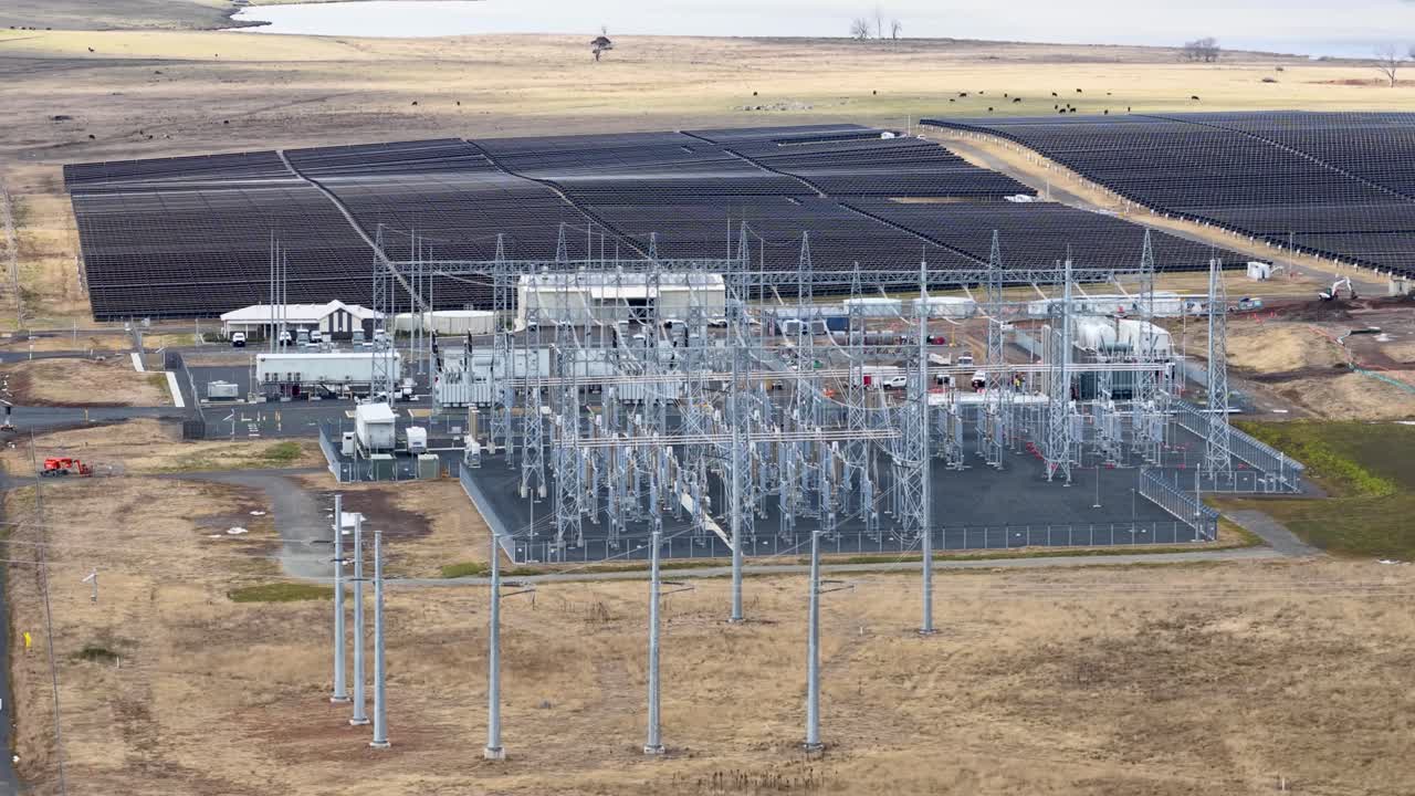 Drone camera smoothly pans across solar farm, substation, and power lines in daylight landscape