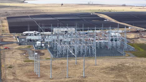 Drone camera smoothly pans across solar farm, substation, and power lines in daylight landscape