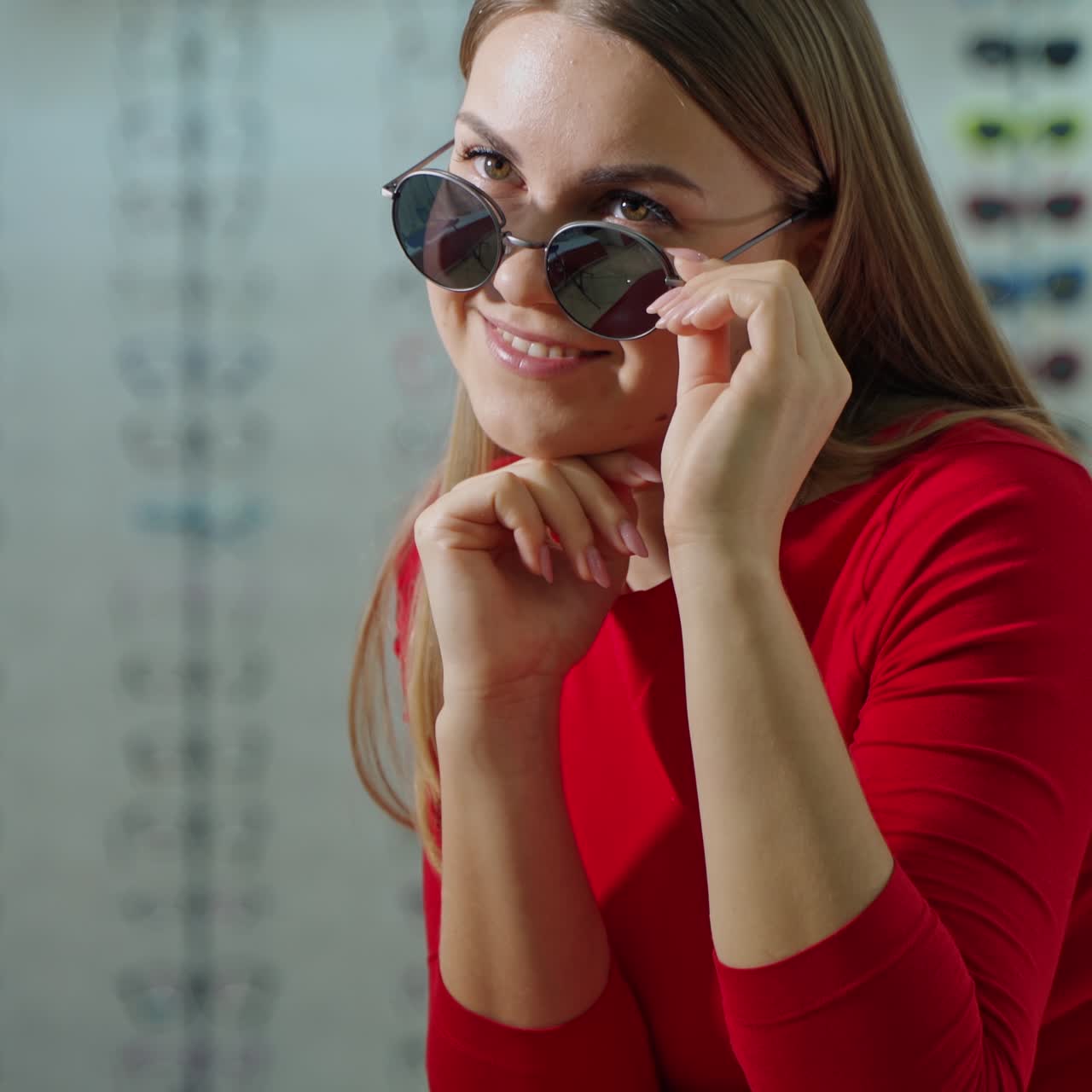 Beautiful girl in trendy sunglasses. Smiling young woman tries on new black glasses on the blurred background of an optical shop.