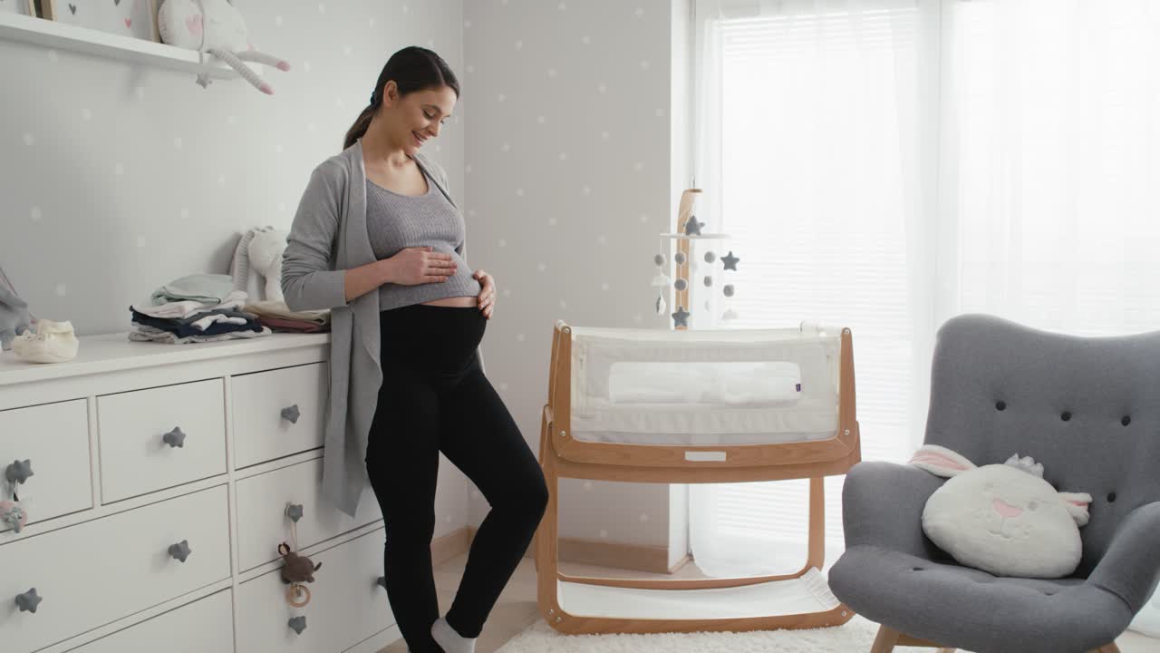 Caucasian woman in advanced pregnancy standing in the baby's room next to the crib.