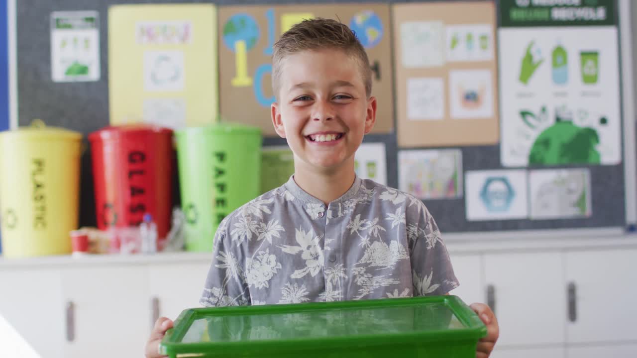 Caucasian schoolboy smiling, holding recycling bin, standing in classroom