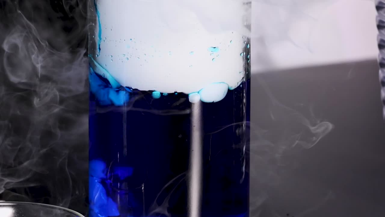 A vibrant blue liquid reacts, creating bubbling and swirling smoke effects in a glass container.
