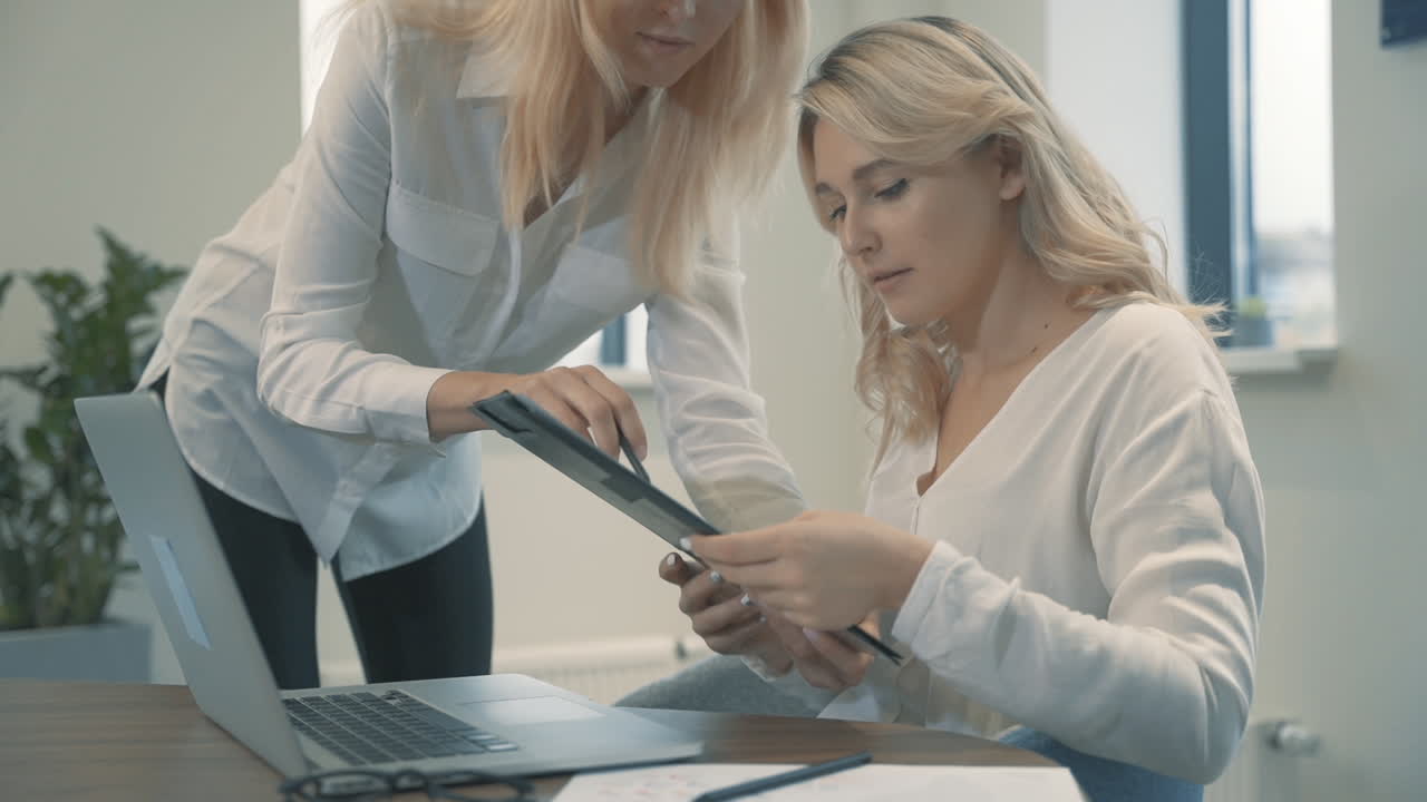 Pretty Young Blonde Woman Works With Laptop, A Coworker Shows Her Some Business Documents