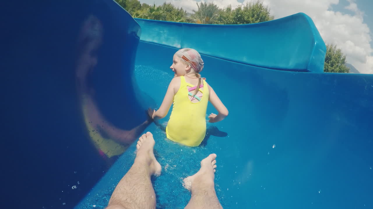 una familia feliz con un niño está montando un tobogán de agua riendo agitando sus manos a la cámara lenta m