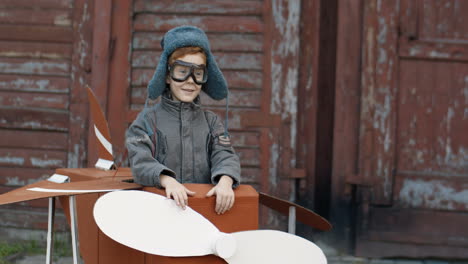 Cute-Little-Red-Haired-Boy-In-Hat-And-Glasses-Walking-Outdoors-In-A-Cardboard-Plane-Dreaming-To-Be-An-Aviator