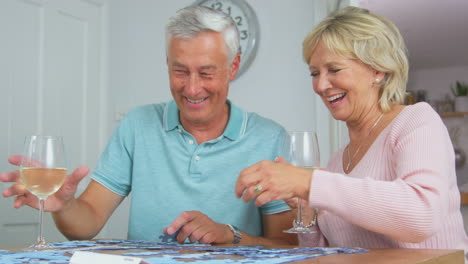 Pareja-De-Jubilados-Mayores-Sentados-A-La-Mesa-En-Casa-Con-Una-Copa-De-Vino-Haciendo-Rompecabezas