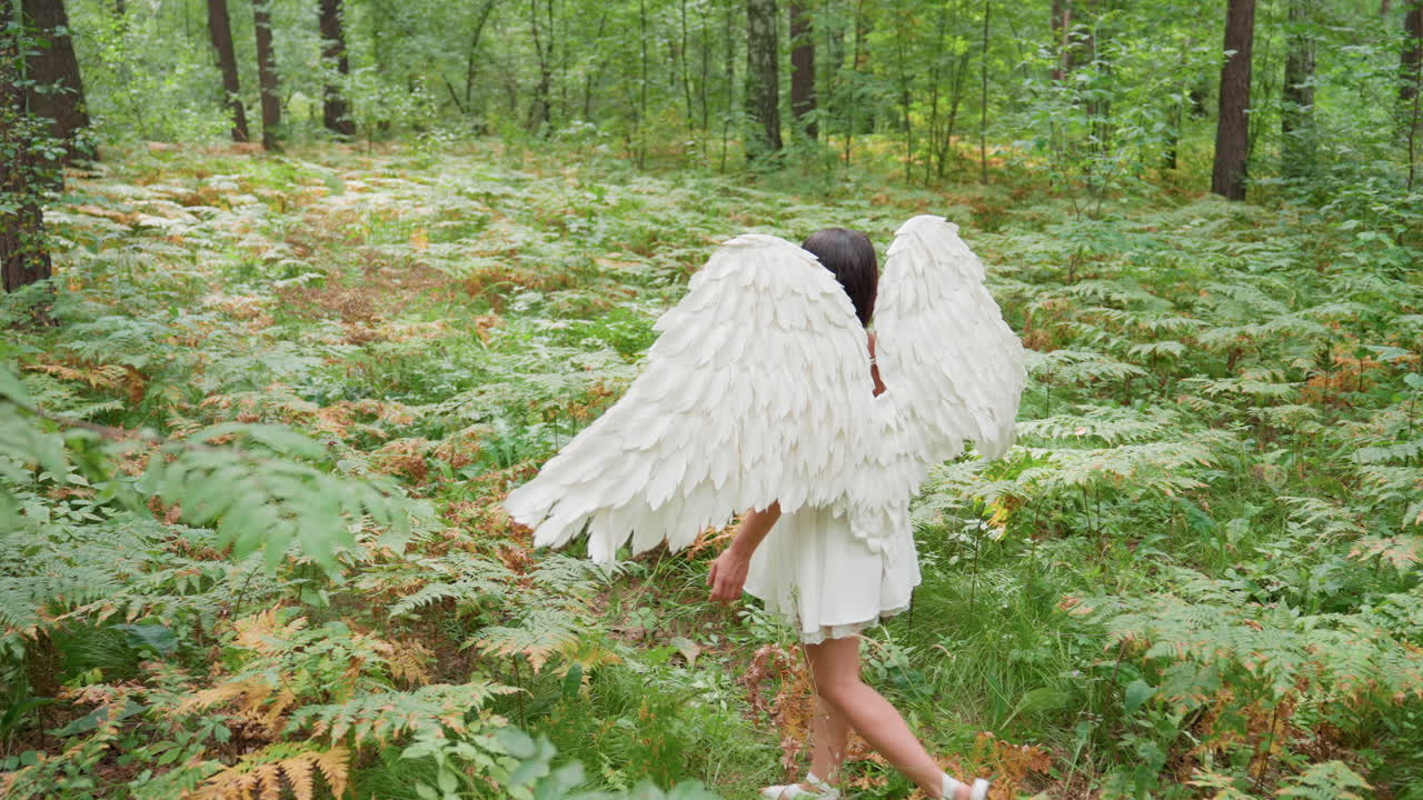 Young lady dressed as mystical forest guardian with white wings walks gently along lush green fern-covered path holding staff, surrounded by vibrant foliage and serene woodland atmosphere