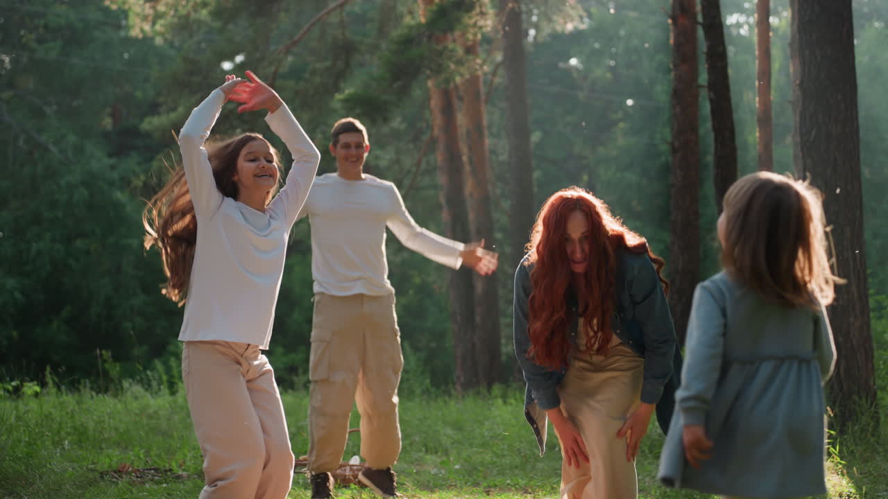 Mom arranging blanket on grass while children jump joyfully with raised hands under glowing sunlight, surrounded by green forest, expressing happiness, playfulness, and family bonding