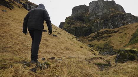 Niedriger-Winkel-Des-Mannes,-Der-Auf-Gelbem-Gras-In-Isländischer-Landschaft-In-Richtung-Einer-Vulkanischen-Klippe-Geht