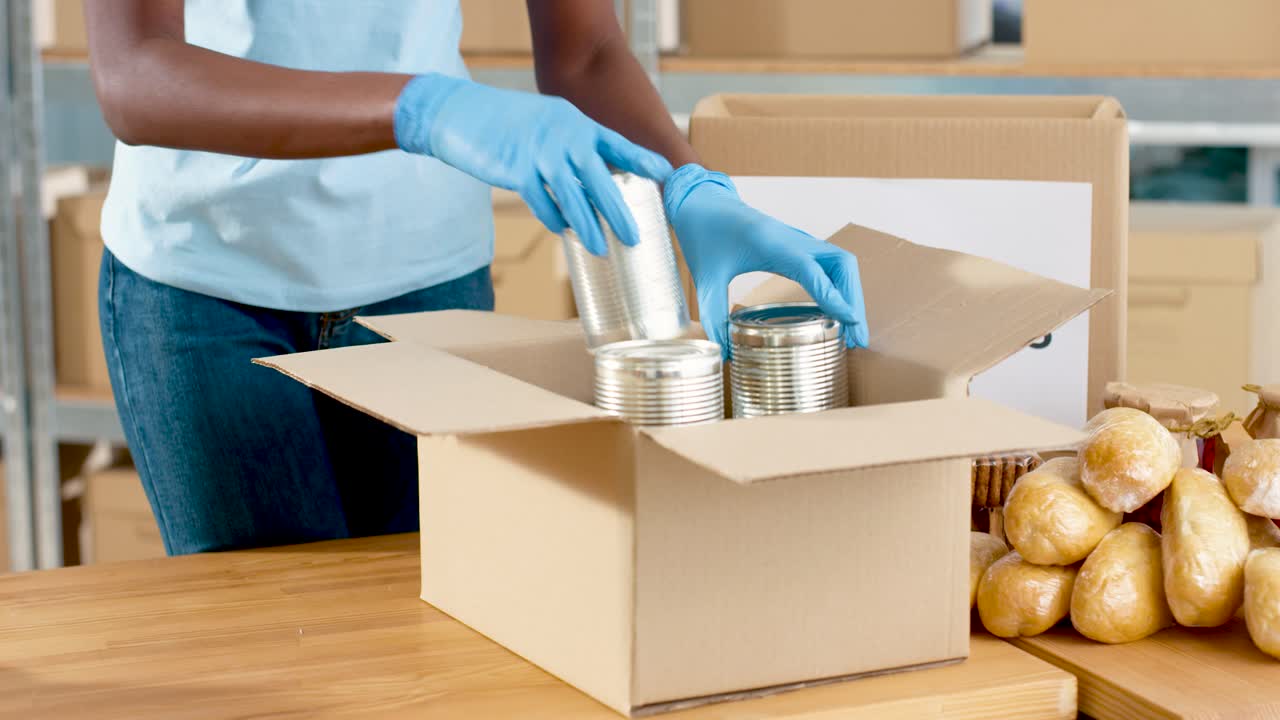 vista cercana de la caja de embalaje de manos voluntarias afroamericanas con latas de conservas en almacén de caridad