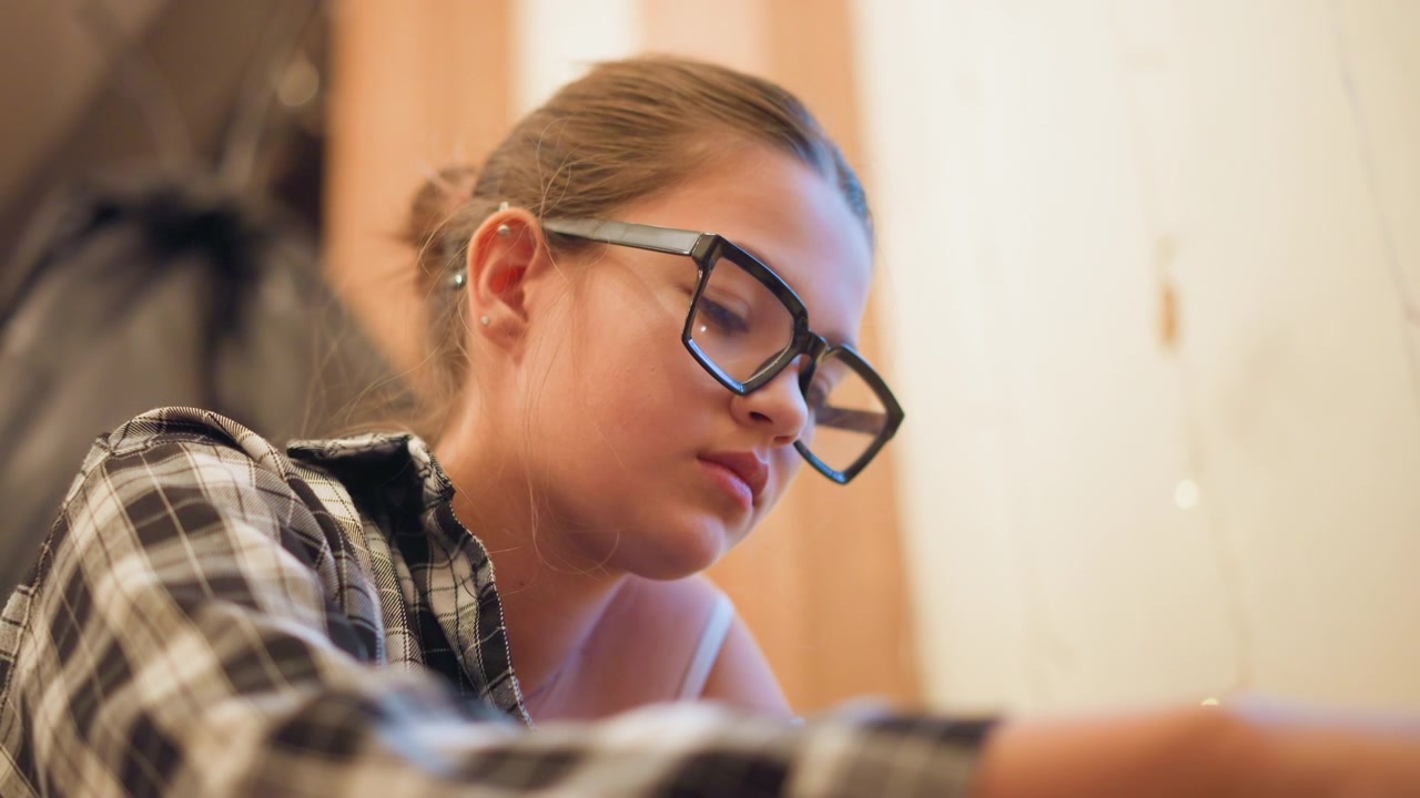 Close up of young woman wearing glasses and plaid shirt focused on unseen task. Her hair is tied back with visible earrings set against soft indoor lighting with warm tones. Background