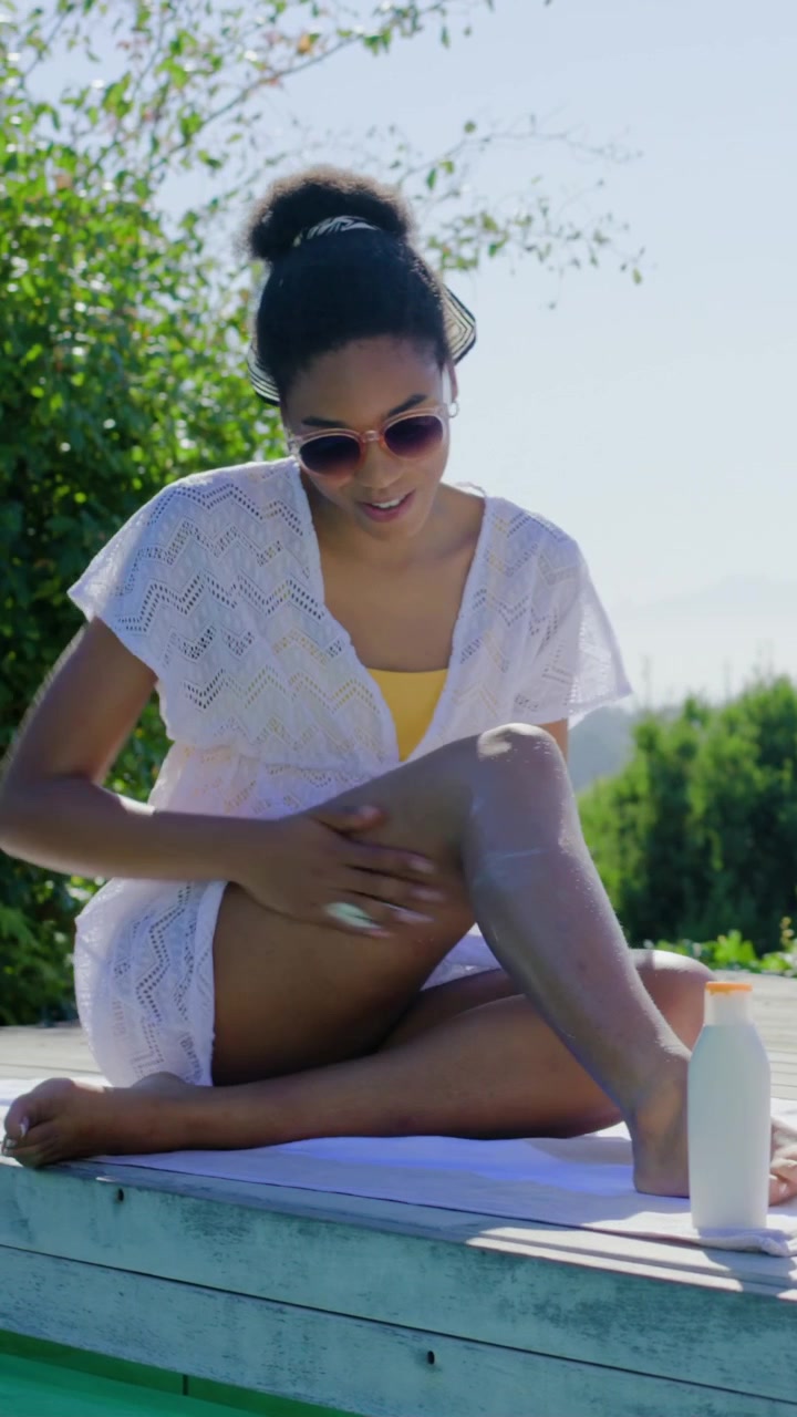 Vertical video: Applying woman in yellow swimsuit protecting legs from sun on deck with sunscreen