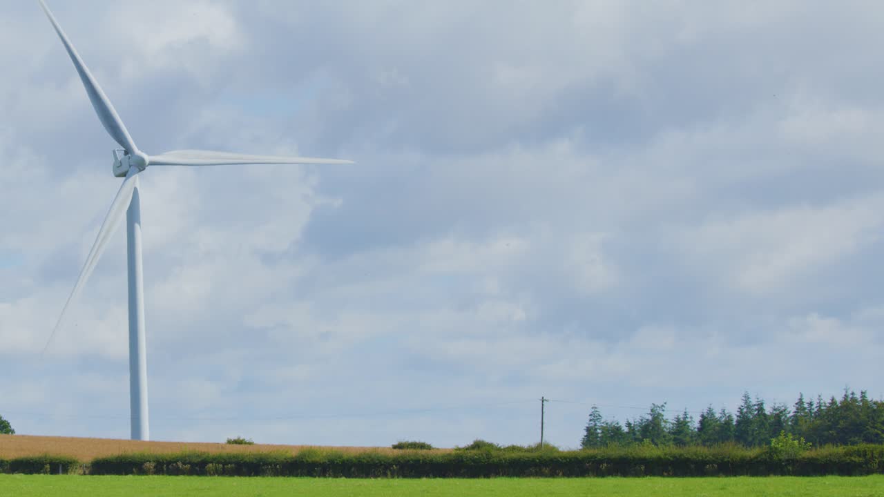 A large wind turbine steadily spins on open farmland bordered by trees, captured in daylight with soft, diffuse natural lighting and a stationary wide shot