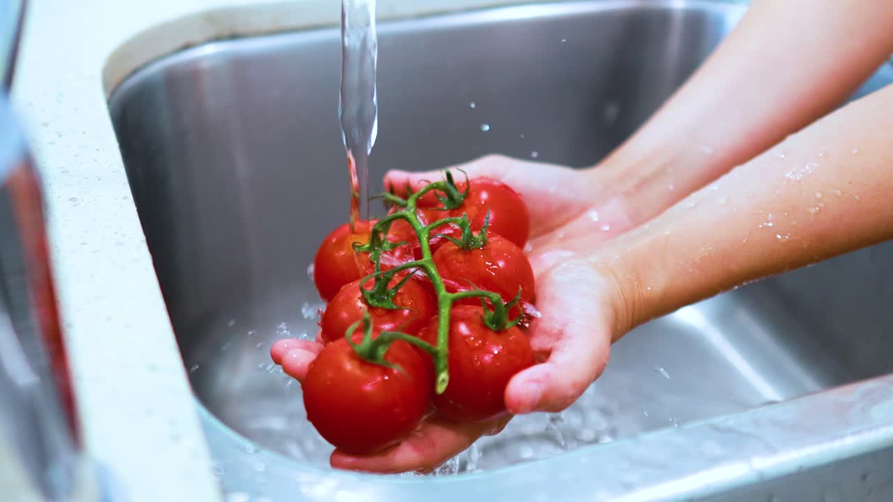 Hands thoroughly wash a cluster of ripe vine tomatoes under running water in a modern kitchen sink, using bright natural lighting and steady camera framing