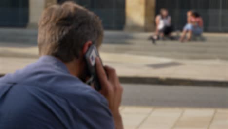 Defokussierte-Aufnahme-Eines-Mannes,-Der-Mit-Seinem-Telefon-Spricht,-Während-Er-Auf-Der-Straße-In-Oxford-Sitzt-02