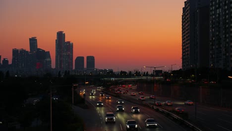 evening traffic with headlights on moves along the gangbyeonbuk-ro expressway in seoul, set against a beautiful orange sunset and the silhouetted skyline of yeouido