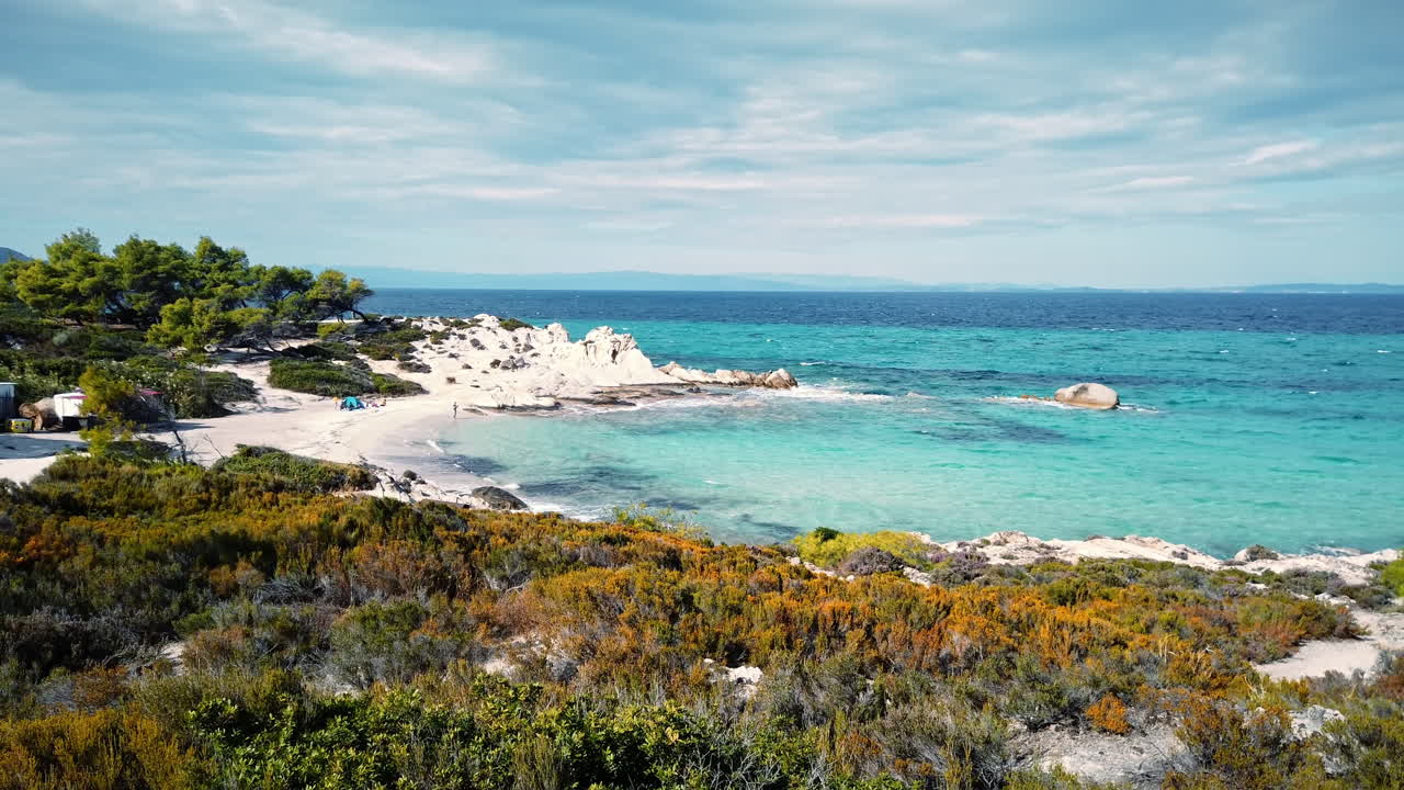 Aegean sea coast with beach. Greenery around, rocks and bushes, blue water with waves, Greece. Slow motion