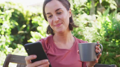 Smiling-caucasian-woman-using-smartphone-and-holding-mug-of-tea-sitting-in-sunny-garden