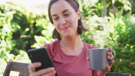 Mujer-Caucásica-Sonriente-Usando-Un-Teléfono-Inteligente-Y-Sosteniendo-Una-Taza-De-Té-Sentada-En-Un-Jardín-Soleado