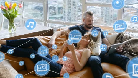 Animation-of-connections-over-caucasian-couple-lying-on-sofa-with-smartphone-and-tablet