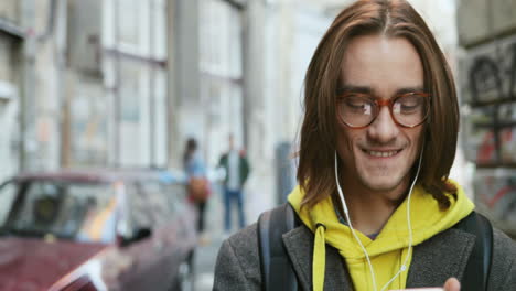 close-up view of caucasian young man in stylish look and headphones smiling and typing on the smartphone in the street