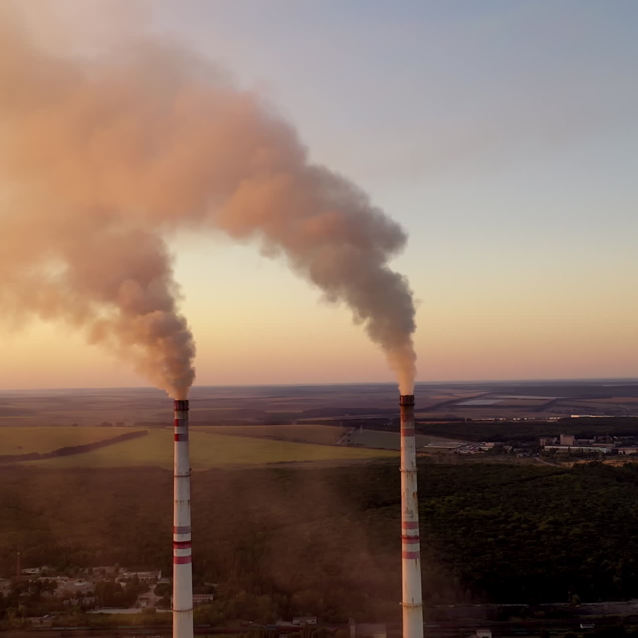 Industrial pipes with smoke. Metallurgical factory among green fields and forests in the evening. Aerial view.