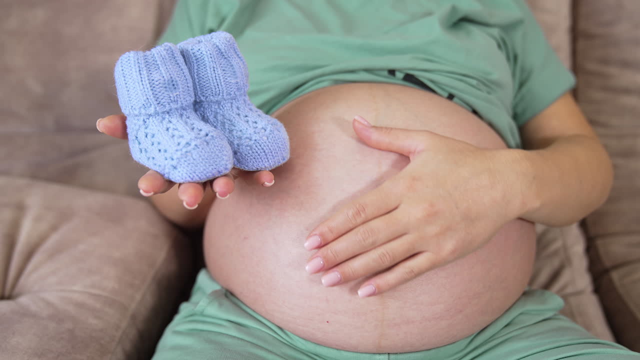 Pregnant woman sitting on the sofa and caressing her big tummy. She's stretching forward her right hand with tiny baby socks on it. Blue baby booties close up.