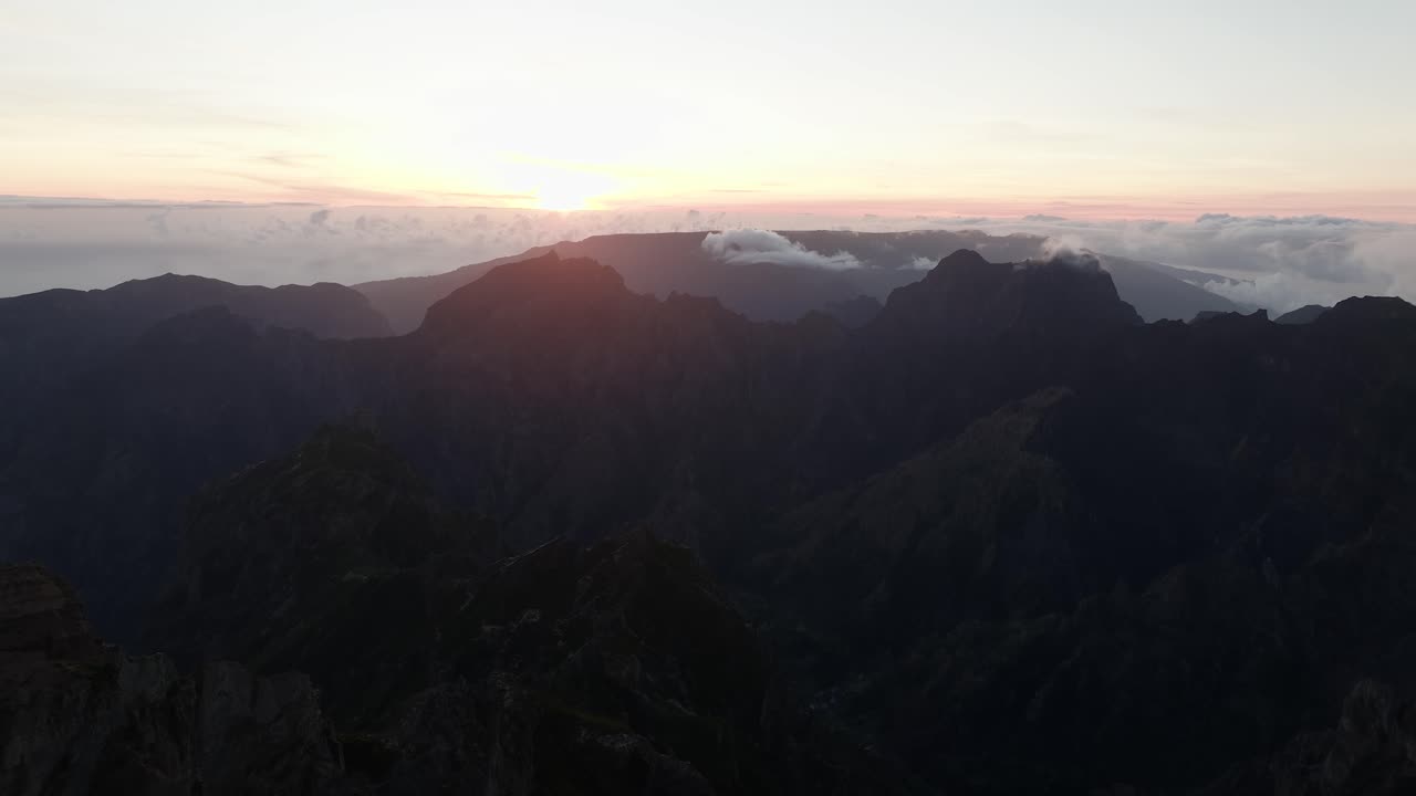 Dramatic aerial of Pico do Arieiro at sunset, with rugged peaks silhouetted against a glowing sky above thick clouds.