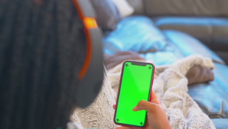 Adolescente-En-El-Sofá-Con-Auriculares-Inalámbricos-Transmitiendo-Contenido-Desde-Un-Teléfono-Móvil-De-Pantalla-Verde