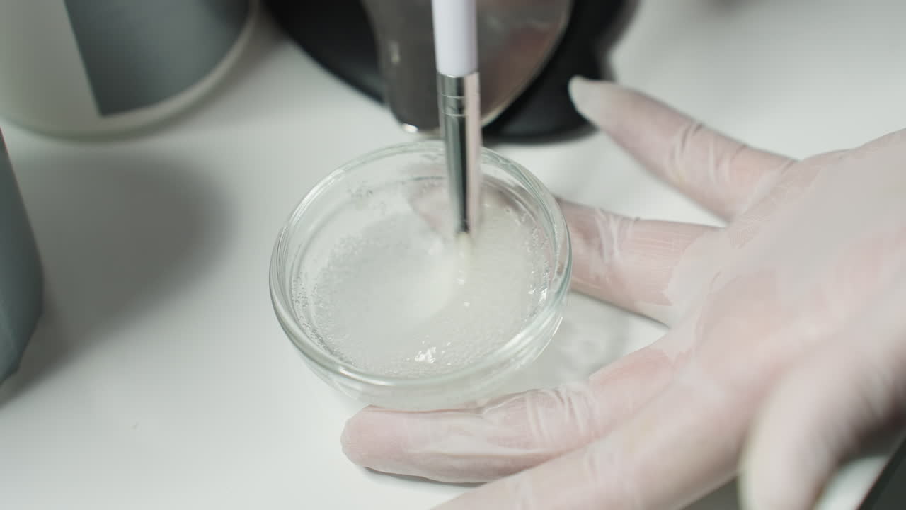 Therapist Hand In Clinical Gloves Stirring Facial Scrub Gel With Bubbles In Clear Glass Bowl On White Tray For Professional Skin Treatment Under Soft Lamp Light In Modern Spa Setting