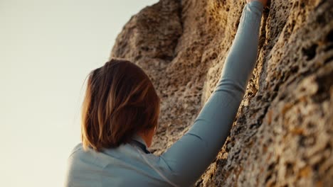 Close-up-shot-of-a-blonde-girl-with-a-bob-hairstyle-in-a-blue-jacket-climbing-a-steep-rock.-Girl-Climber-climbs-up-the-rock-and-looks-for-the-right-places-to-place-her-fingers-for-easy-climbing-on-the-rock