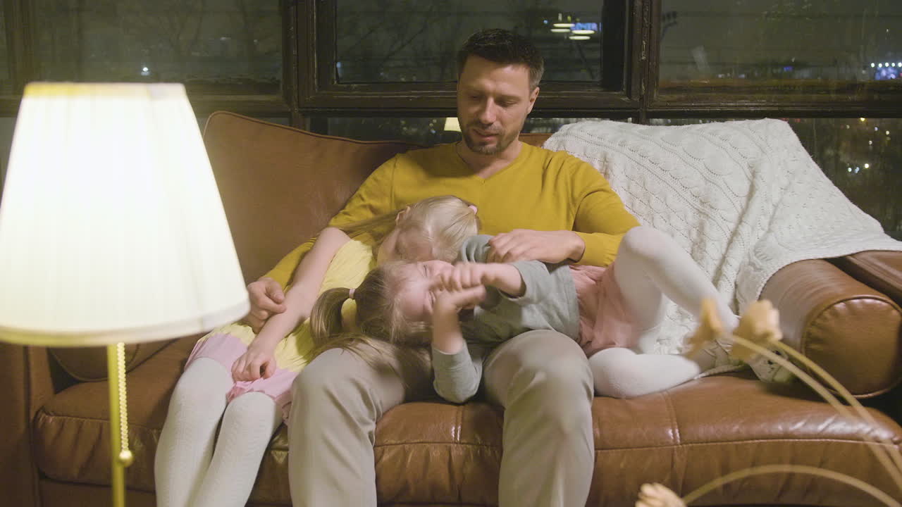 Happy Father Sitting On Sofa With His Two Sleepy Little Girls Who Leaning Their Heads On His Lap