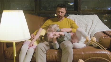 happy father sitting on sofa with his two sleepy little girls who leaning their heads on his lap