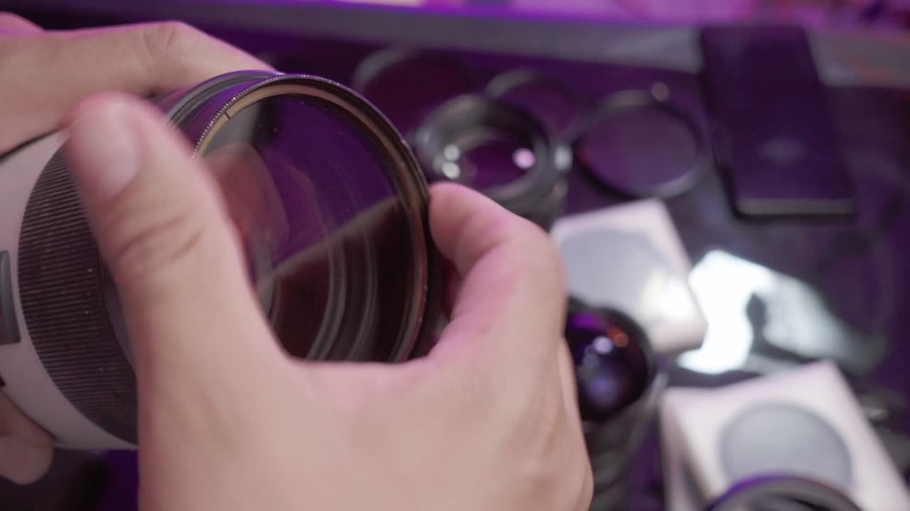 Person holding a camera lens examines it closely with various lenses and accessories around