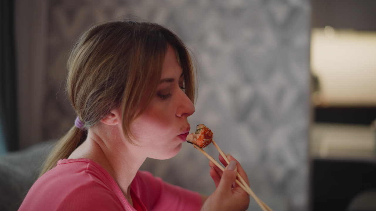 Lady in pink polo shirt seated on couch savoring sushi with chopstick in cozy indoor setting featuring soft blur background peaceful expression and gentle lighting