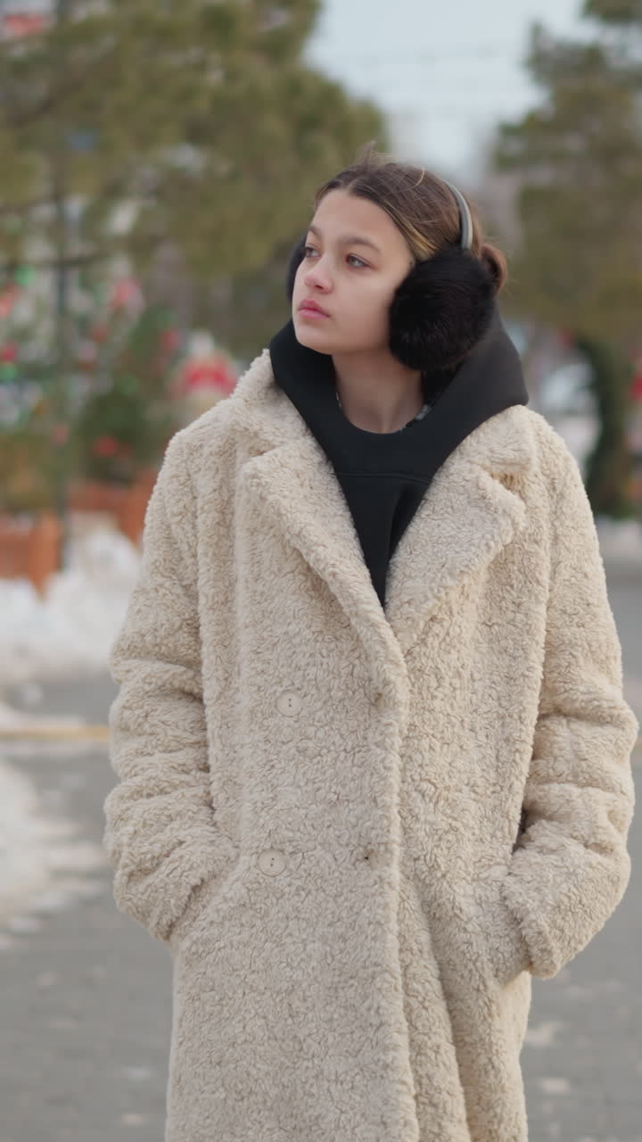 베이지색 시어링 코트를 입고 검은색 귀마개와 후드 티셔츠를 착용한 젊은 여성이 눈 덮인 인도를 걸으며 주머니에 손을 넣고 생각에 잠긴 시선으로 부드럽고 은은한 빛을 받으며 도시의 나무들이 흐릿하게 보이는 배경 속에서 아늑한 겨울을 보내고 있어요.