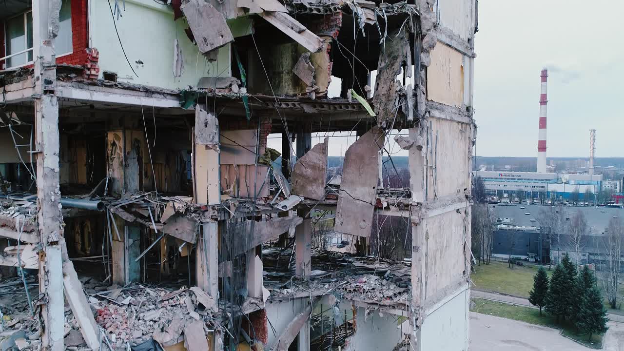 Drone ascends showing damaged Soviet-era block and Riga industrial skyline