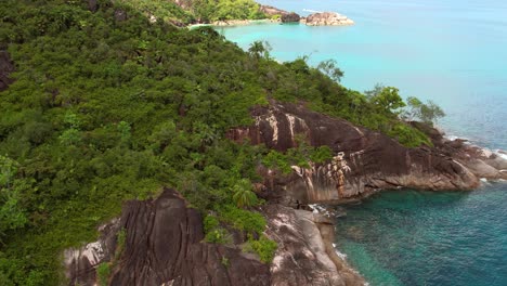 drone shot passing over lush mountain with granite stone and reveal of beach, mahe, seychelles 60 fps