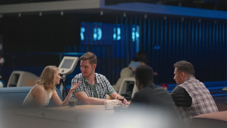 a multi ethnic group of friends in a bowling club are chatting at the table and smiling rejoicing and high-fiving a friend who knocked out a strike