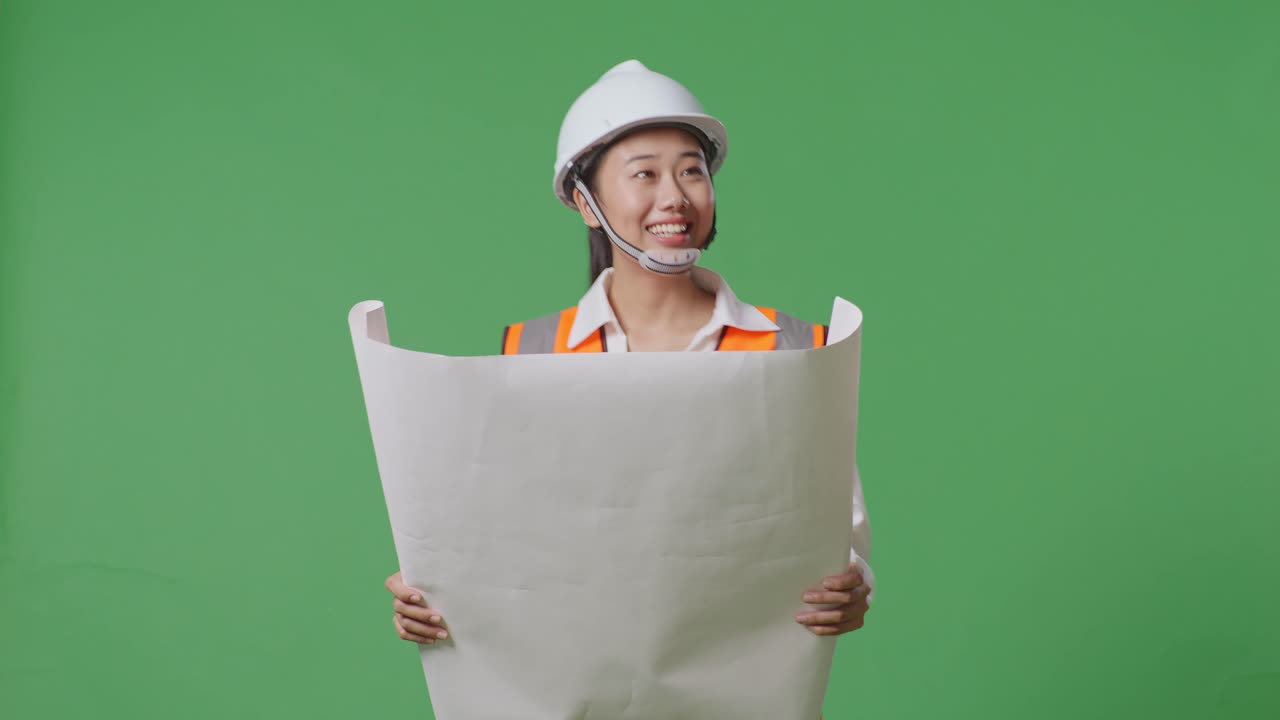 안전 헬을 입은 아시아 여성 엔지니어가 손에 청사진을 들고 녹색 스크린 배경 스튜디오에서 는 동안 주변을 둘러보고 있습니다.