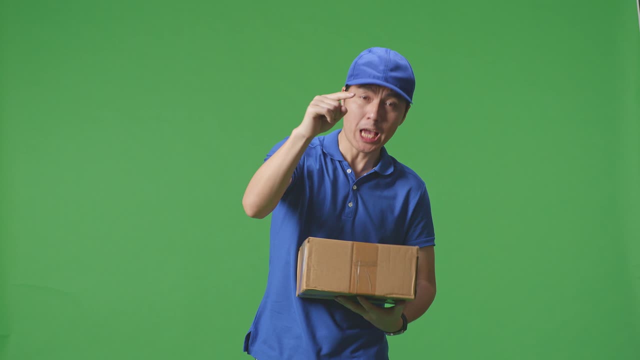 푸른 유니폼을 입은 불만족한 아시아 남성 택배기사가 스튜디오에서 녹색 스크린 배경에 상자를 배달하는 동안 카메라에 소리치고 있습니다.