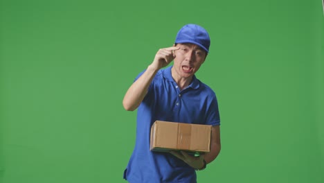 mensajero masculino asiático insatisfecho en uniforme azul gritando a la cámara mientras entrega una caja de cartón en el fondo de la pantalla verde en el estudio