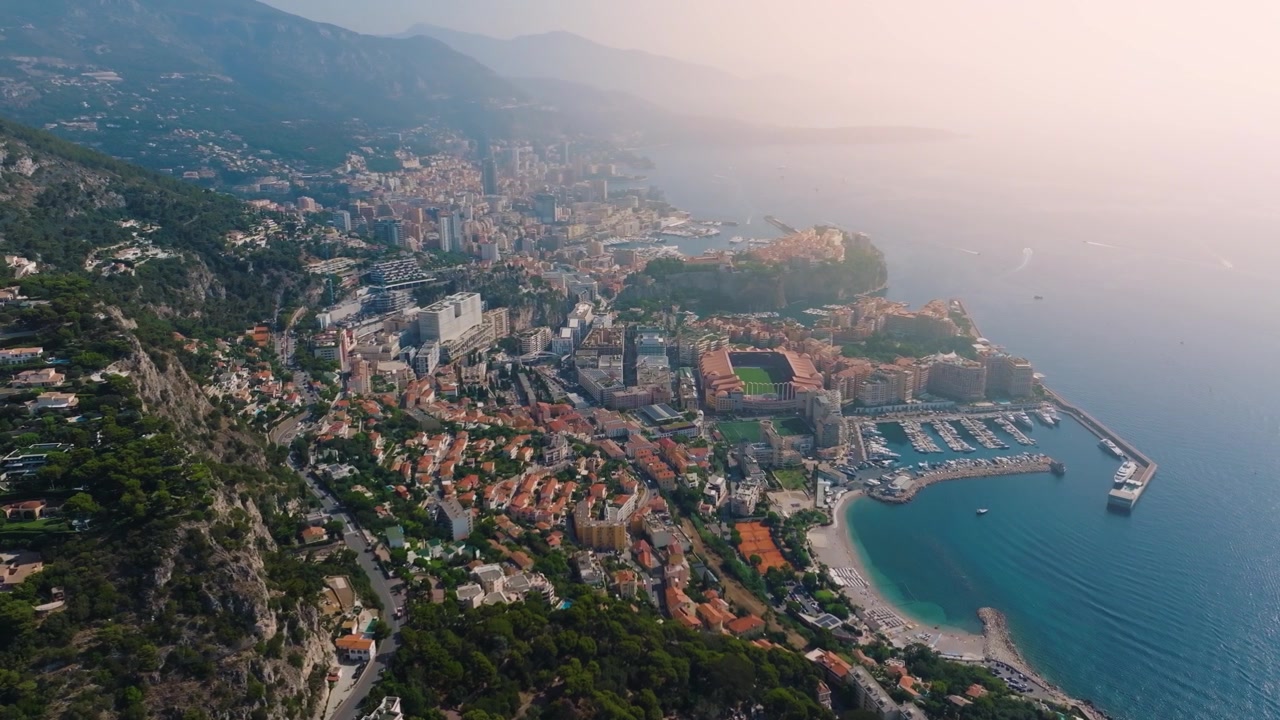 Monaco presenta la costa e gli edifici di lusso mentre il sole tramonta sul Mar Mediterraneo. La vista cattura le barche nel porto e lo skyline della città, mettendo in mostra la sua famosa architettura
