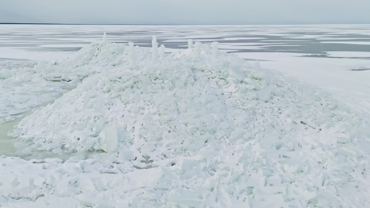 Vista de dron acercándose a crestas de presión cubiertas de nieve y losas de hielo apiladas en un paisaje marino invernal, con patrones de hielo en capas que se extienden hasta el horizonte.
