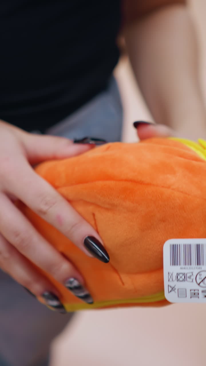 밝게 조명 된 쇼핑몰에서 녹색 세부 사항으로 활기찬 오렌지색 플러시 지갑을 들고 조사하는 여성의 클로즈업, 그녀는 다양한 물건으로 가득 찬 상점 선반 근처에 서서 부드러운 직물을 검사합니다.